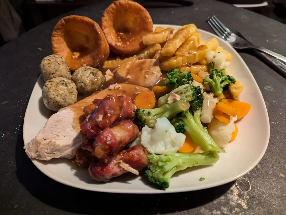 A plate of food featuring roasted chicken, bacon-wrapped sausages, Yorkshire puddings, fried chips, broccoli, cauliflower…