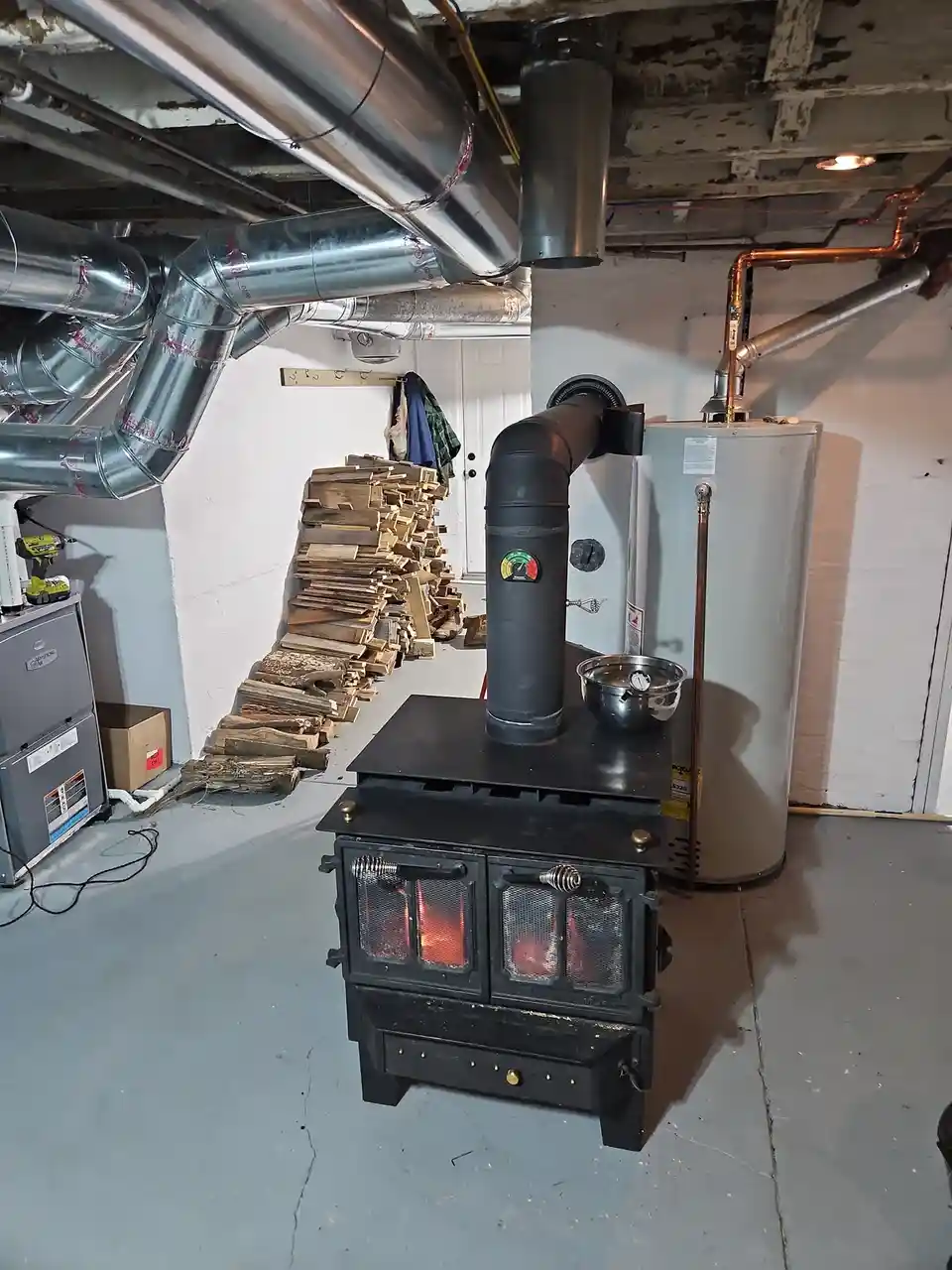 A basement room contains a lit black wood stove with a large stack of firewood beside it, alongside a water heater and HVAC…