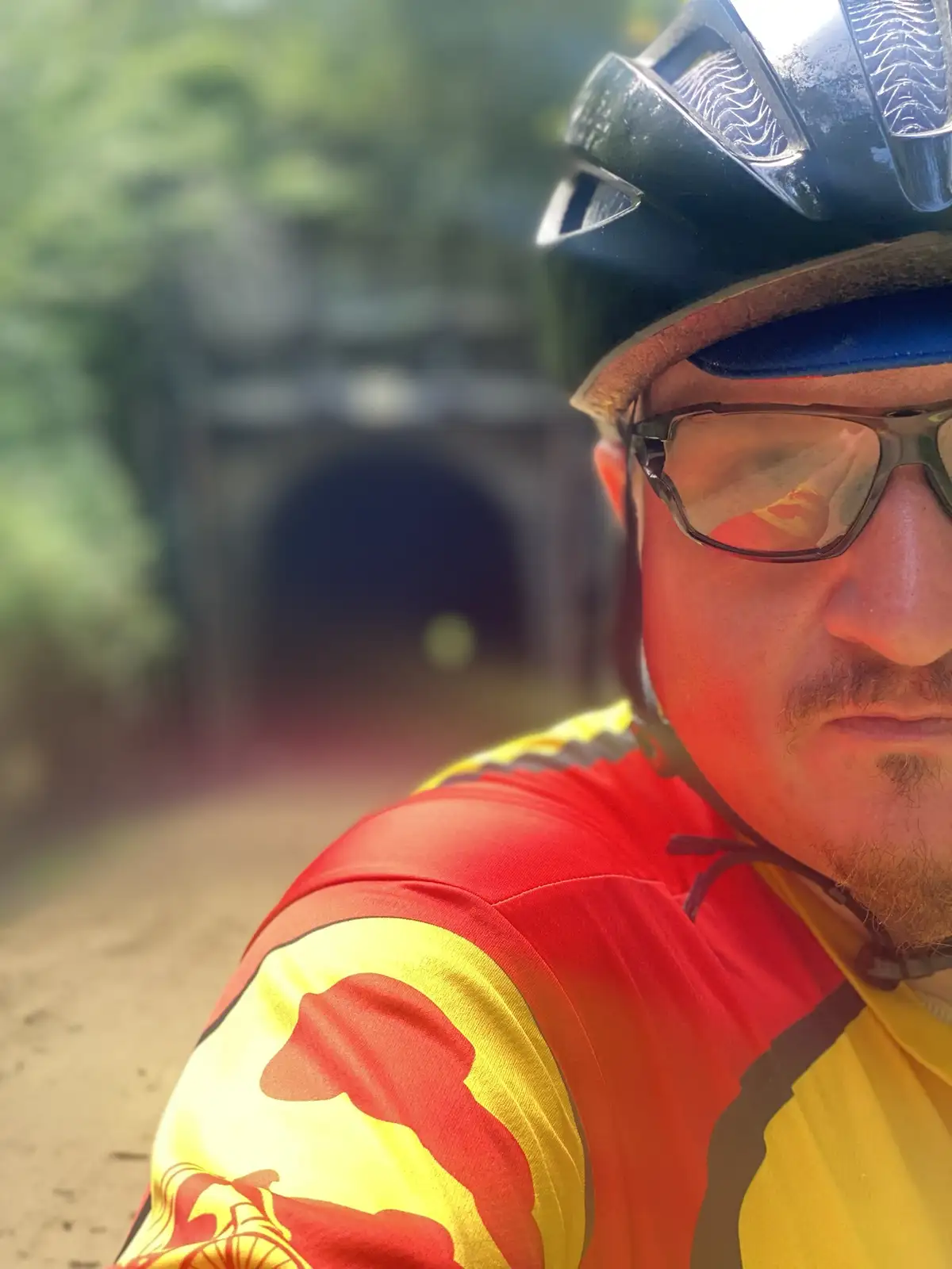 A close-up selfie of a man wearing a black bicycle helmet and sunglasses, looking toward the camera with a neutral…