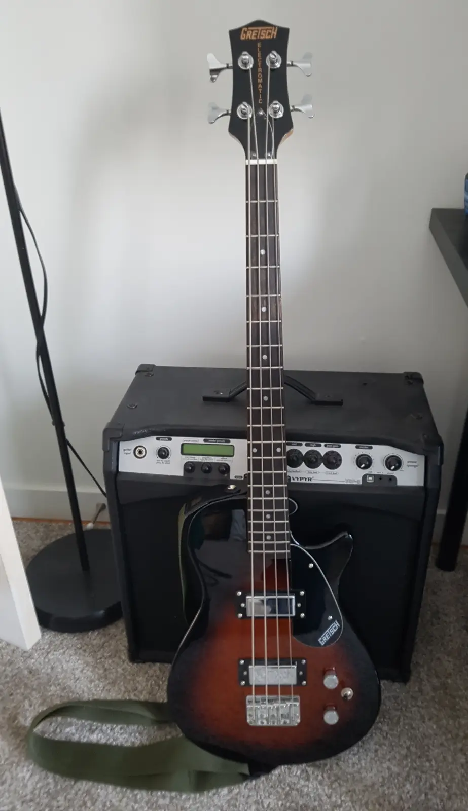 A sunburst-finish Gretsch Electromatic bass guitar stands upright on a carpeted floor, leaning against a black Vypyr bass…