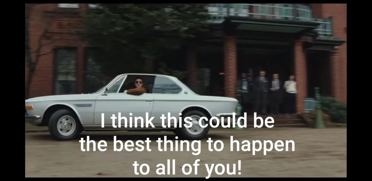 A man in a white vintage car drives past a brick building where several people stand on the porch. He leans out the window…