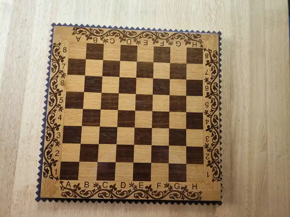 A wooden chessboard with alternating light and dark squares, decorated with ornate floral carvings along its edges and…