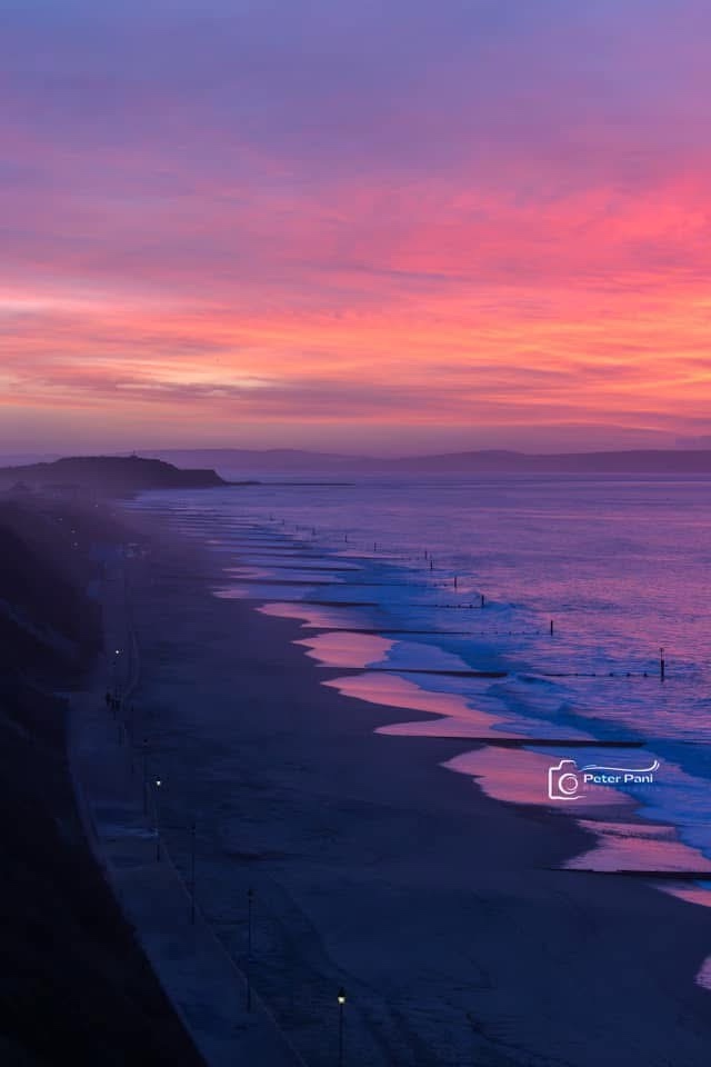 A serene beach at dusk with vibrant pink and purple skies reflected in the ocean waves. A paved walkway with streetlights…