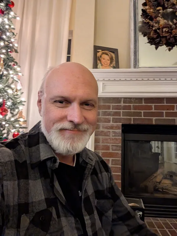 a bald man with a white goatee and a plaid shirt smiles smugly at the camera while sitting in front of a fireplace. Behind…