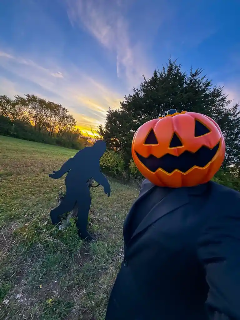 A person wearing a large orange jack-o'-lantern head and a black suit stands in a grassy field at sunset, facing a black…