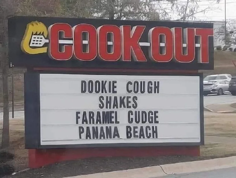 An outdoor sign for the fast-food chain Cook-Out features a marquee board with misspelled menu items: 'DOOKIE COUGH SHAKES'…