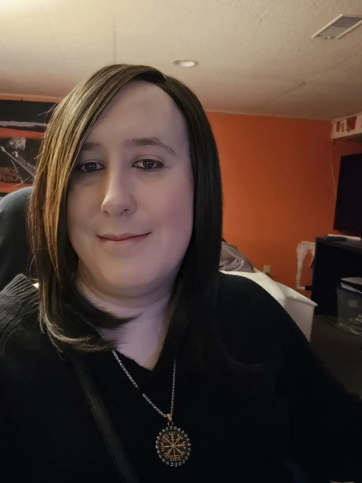 A person with shoulder-length brown hair and a black top smiles faintly while wearing a necklace with a circular pendant…