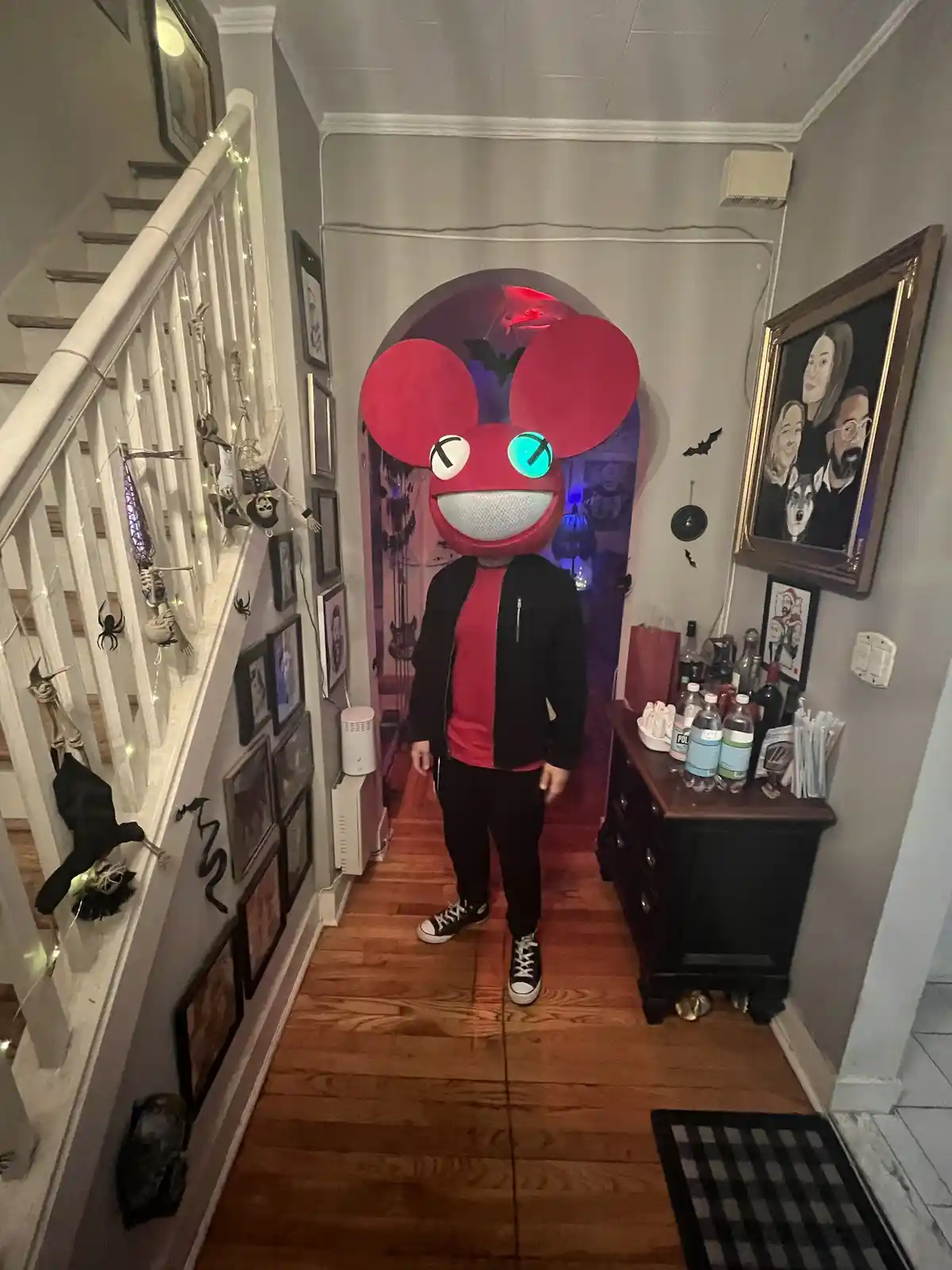 A person stands in a narrow hallway wearing a large, red, cartoonish mouse head costume with one glowing blue eye and one…