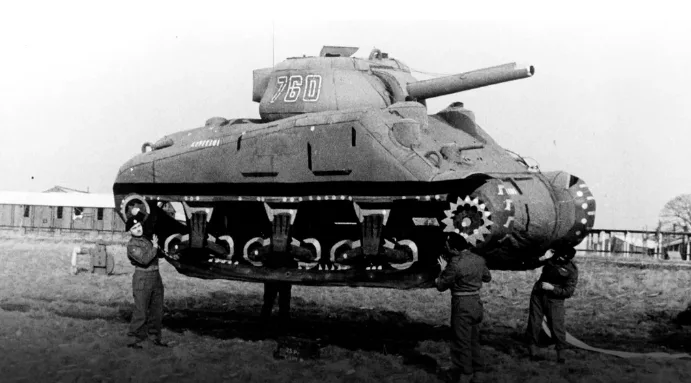 A black-and-white historical photo shows three soldiers standing beneath a massive, oversized inflatable tank replica marked…