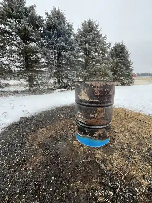 A rusty, weathered metal barrel sits outdoors on a patch of dirt and snow, with frost-covered evergreen trees in the…