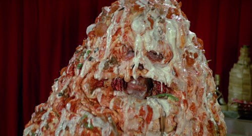 A close-up of a large, messy, and grotesque food creation resembling a human face, covered in sauce and cheese, with a red…