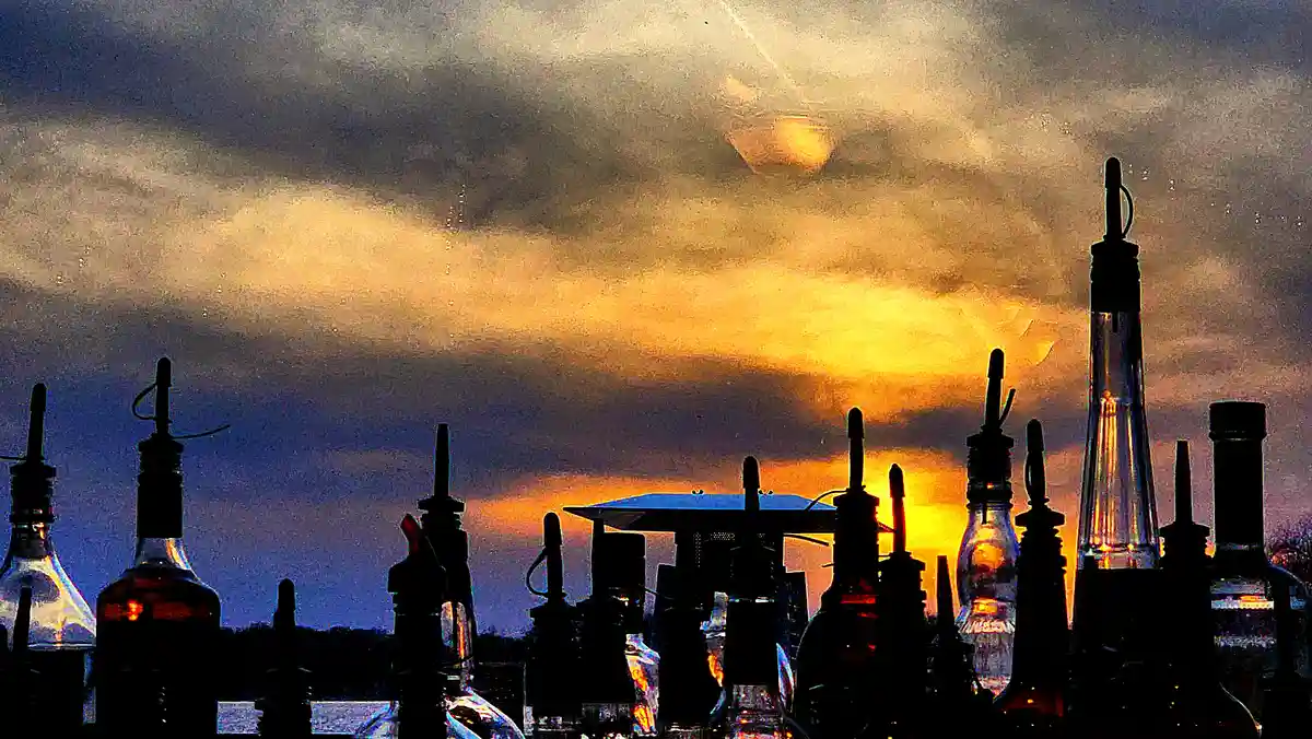 The image shows the silhouettes of numerous glass bottles with pour spouts against a dramatic sunset sky with orange and…