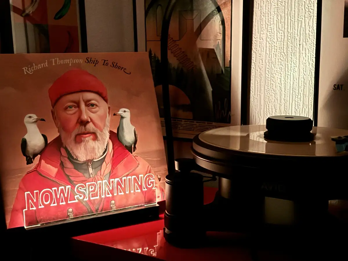 A vinyl record player sits next to a glowing album cover for Richard Thompson's 'Ship To Shore', which features a man in a…