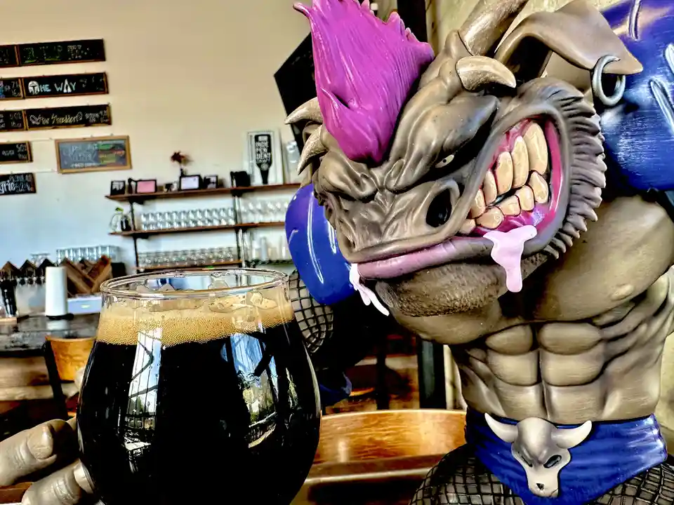 A large, muscular, fantasy-style statue with a demonic face, purple mohawk, and blue armor is holding a glass of dark beer…