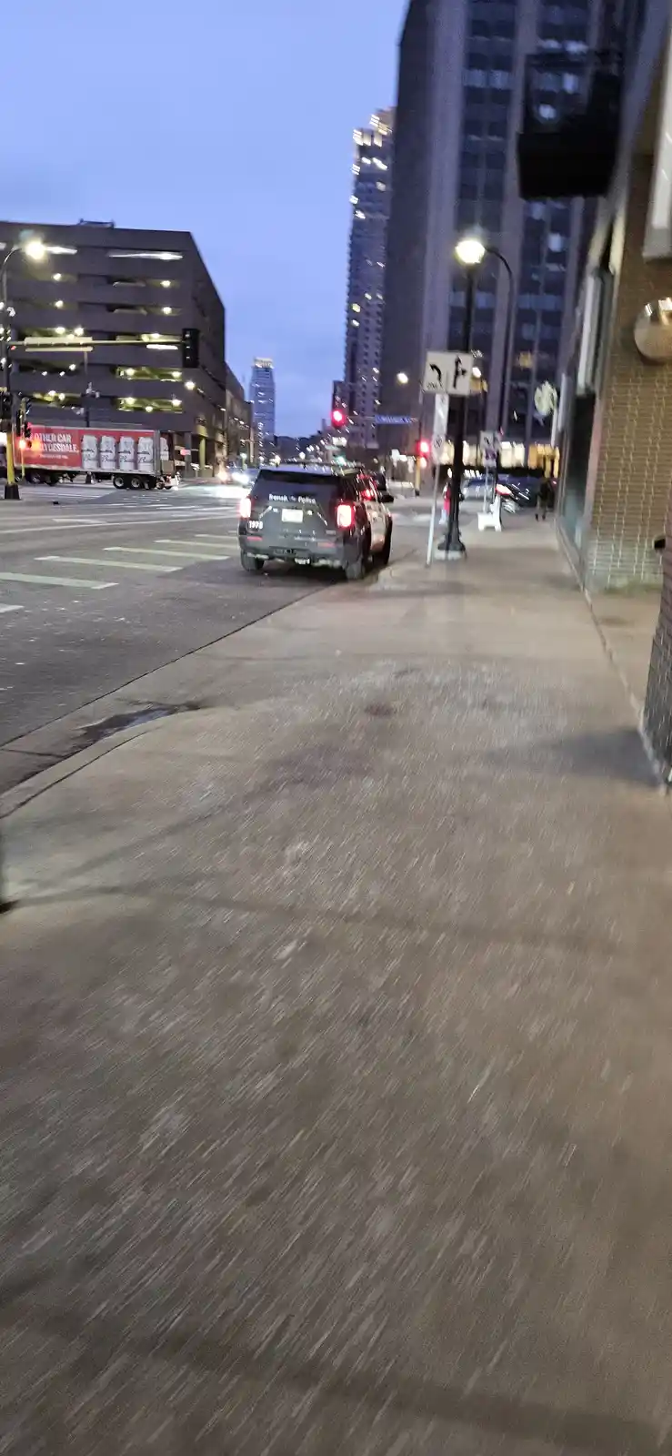 A blurry, low-angle photo of a city sidewalk at dusk, with a dark police SUV stopped at a street intersection. The image has…