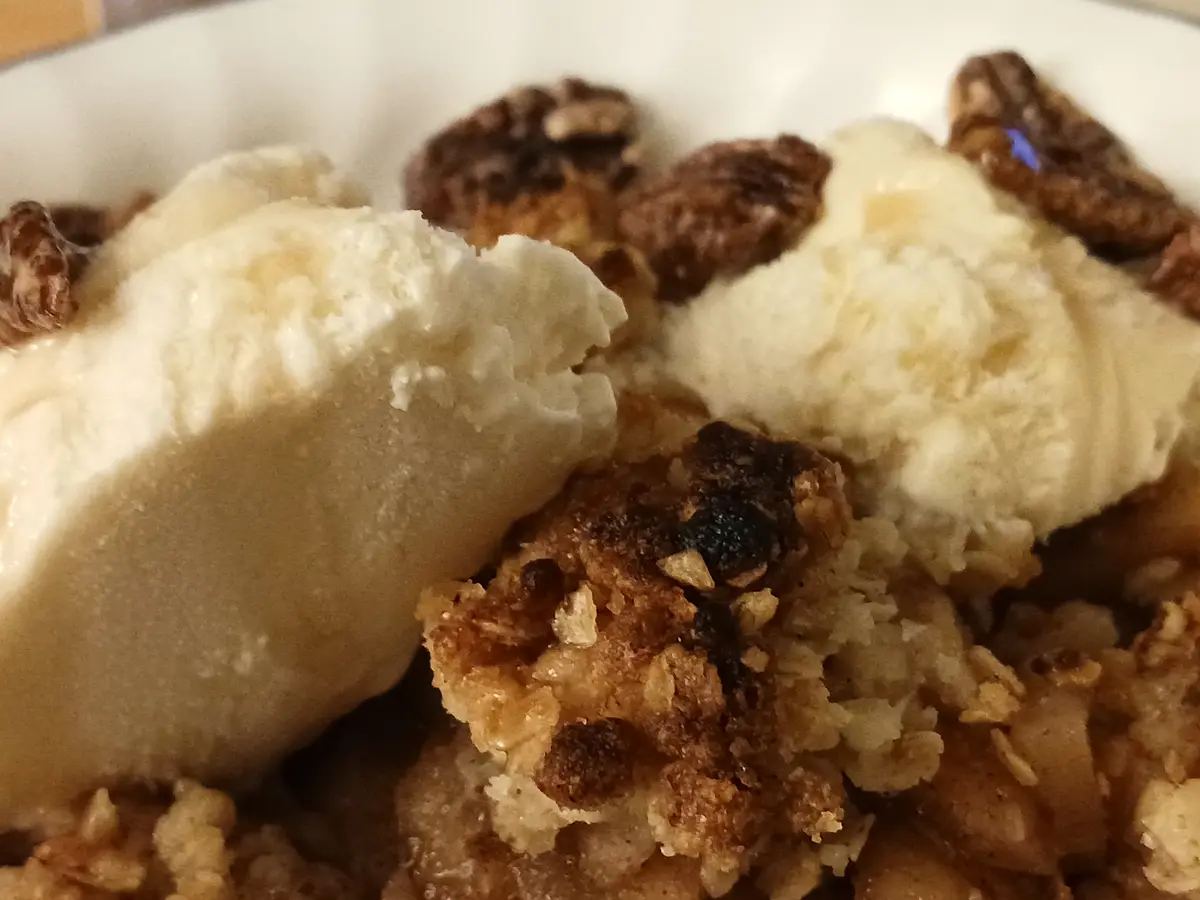 A close-up photo shows a dessert in a white bowl, featuring two scoops of vanilla ice cream melting slightly atop a crumbly…