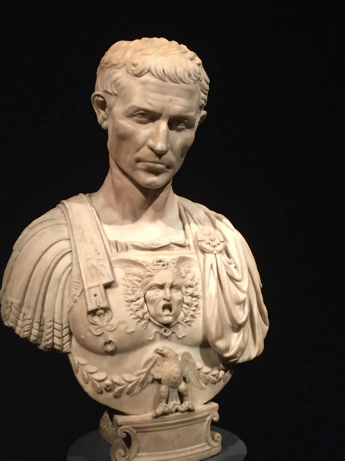 A marble bust of a Roman emperor, likely Augustus, with detailed armor featuring a screaming face and an eagle, set against…