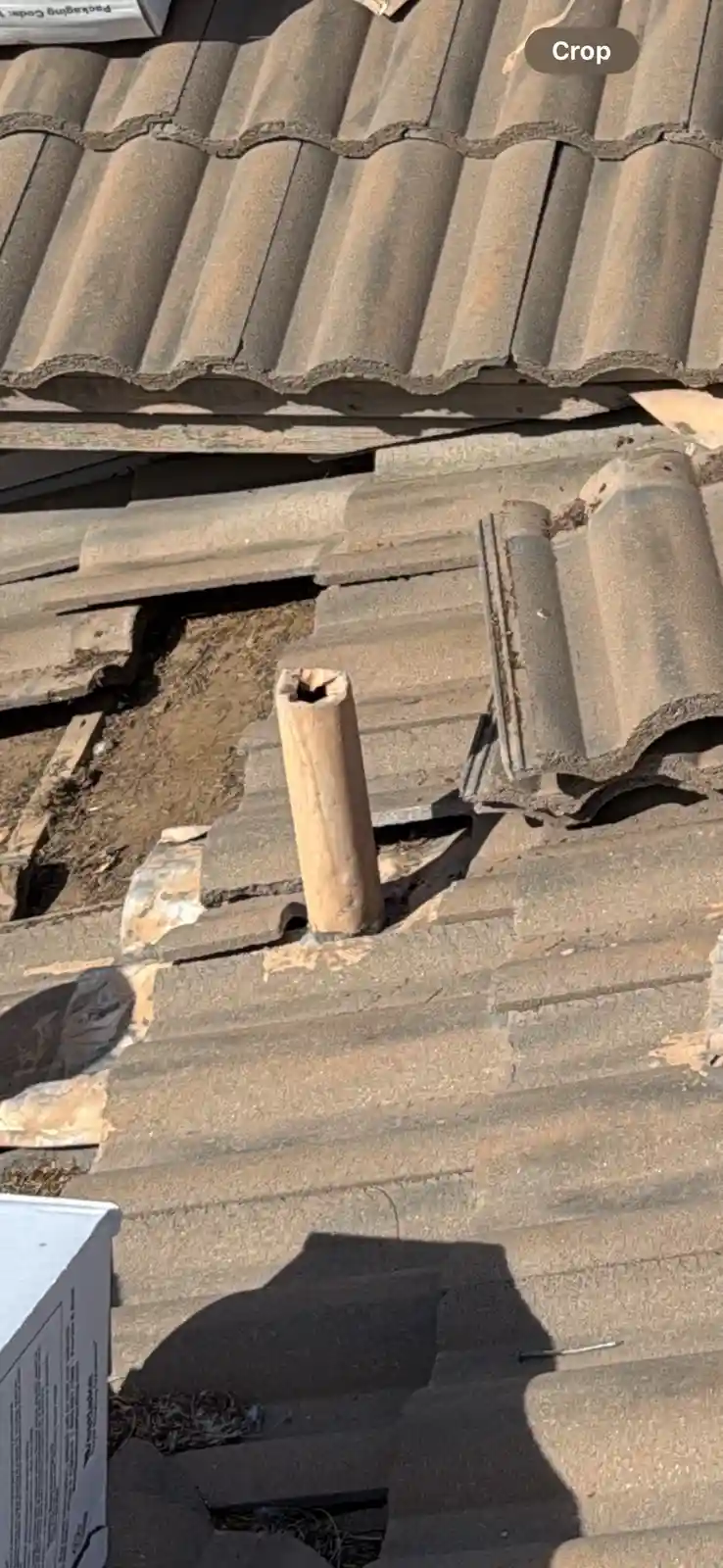 A close-up photo of a damaged roof with broken, dislodged tiles and exposed wood. A short, hollow, cylindrical wooden pipe…