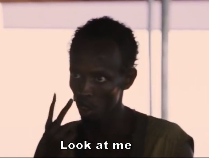 A man with dark skin and short hair stares intensely at the camera while holding up two fingers in a peace sign gesture. The…