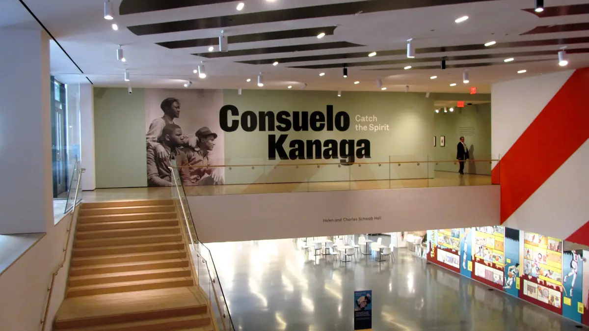 An interior shot of a modern museum or gallery with a wooden staircase leading up to a mezzanine. On the upper level, a…