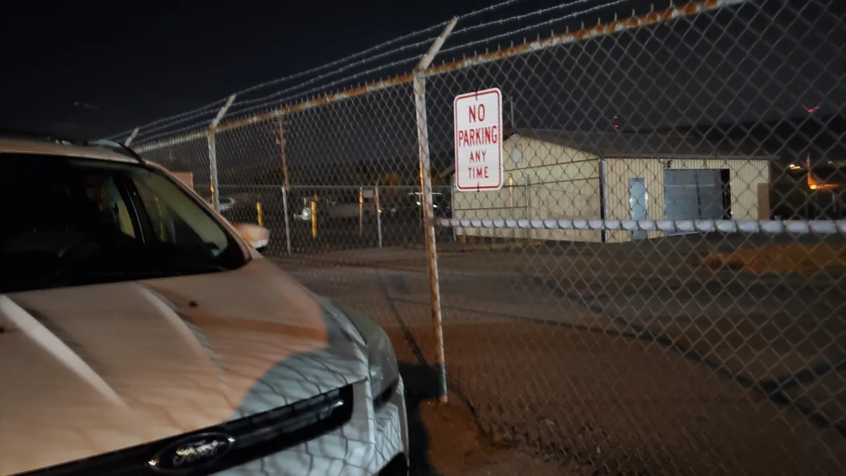 A silver Ford car is parked directly next to a chain-link fence at night, ignoring a red and white 'NO PARKING ANY TIME'…