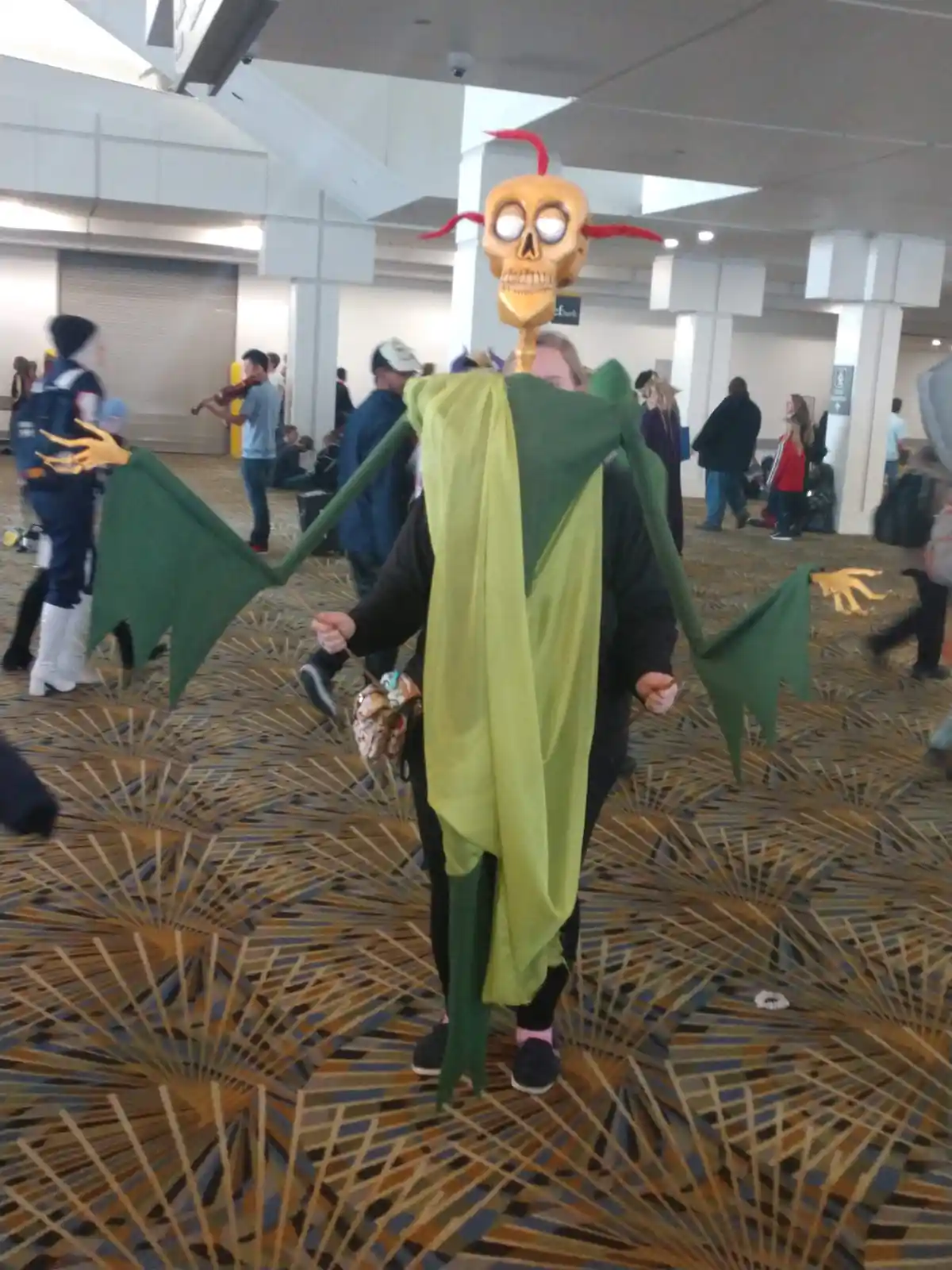 A person in a convention hall is wearing a full-body costume of a green, skeletal dragon with large wings, a skull head with…