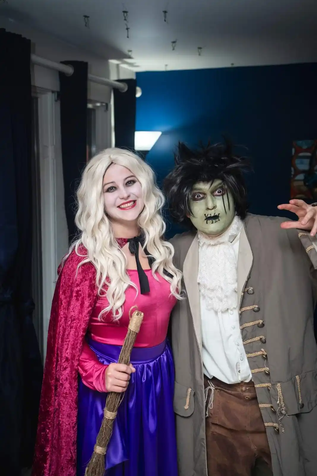 Two people dressed in Halloween costumes pose together; one is a smiling woman in a pink and purple witch outfit with a…