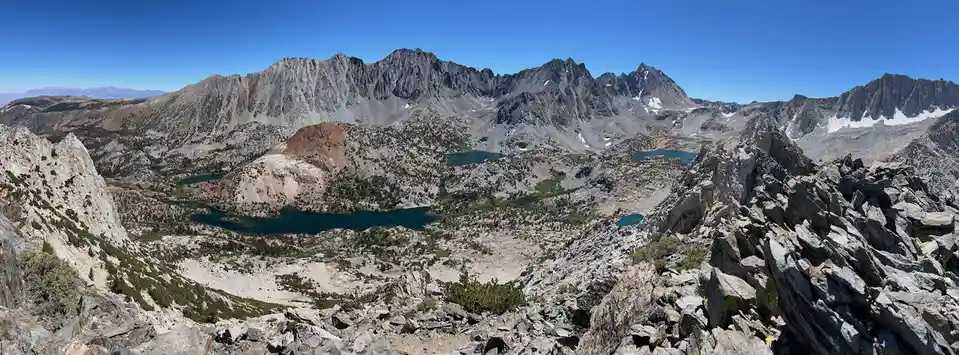 A panoramic photo of a rugged mountain landscape with jagged peaks, rocky terrain, and several alpine lakes under a clear…