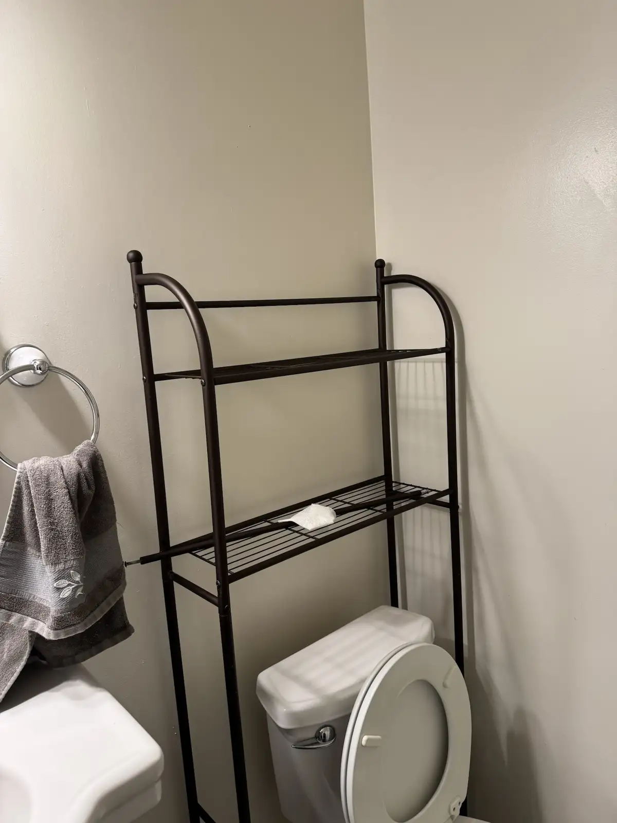A bathroom scene shows a dark metal over-the-toilet shelf with a single white tissue resting on its middle wire rack. To the…