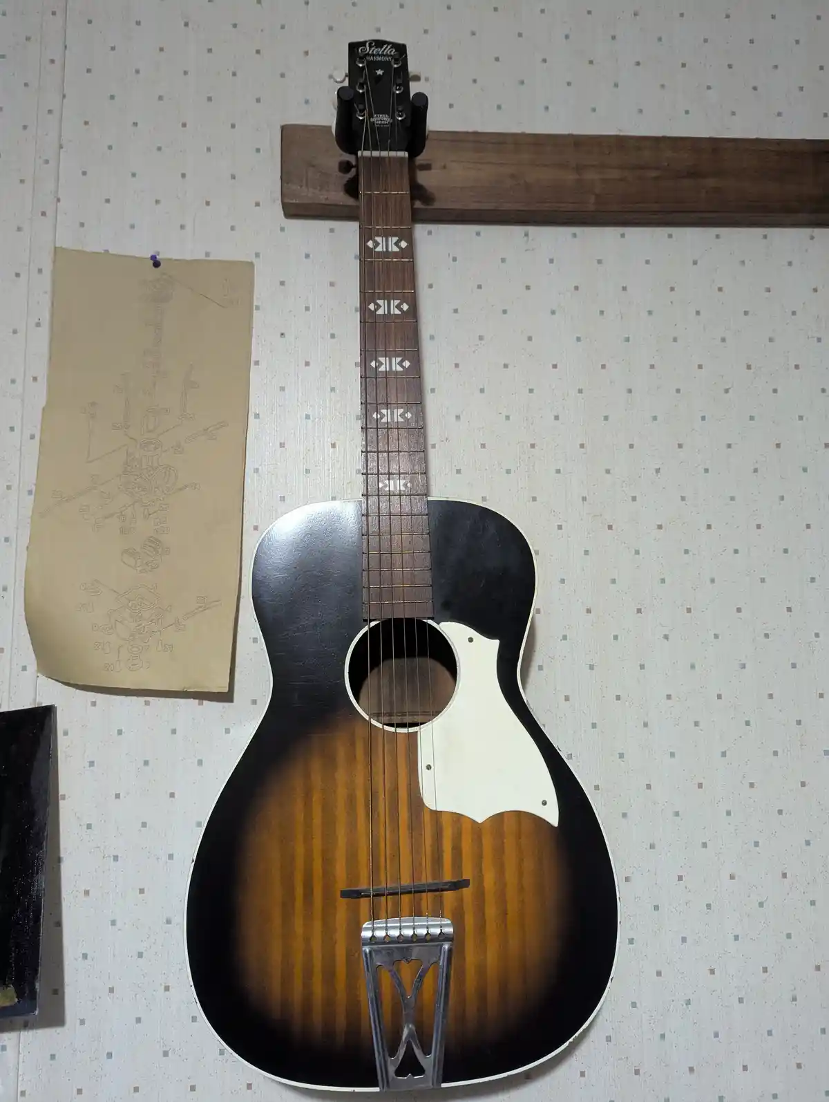 An acoustic guitar with a sunburst finish and a metal bridge is mounted vertically on a wooden rack against patterned…