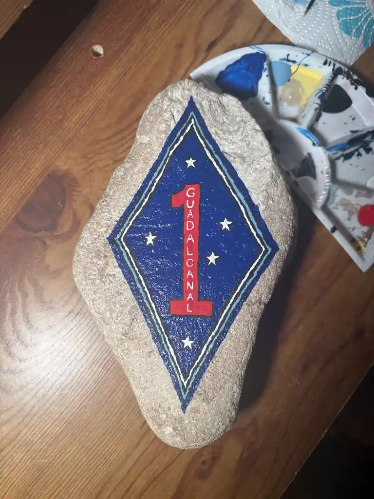 A hand-painted rock on a wooden table features a blue diamond-shaped insignia with a red number '1' and the word…
