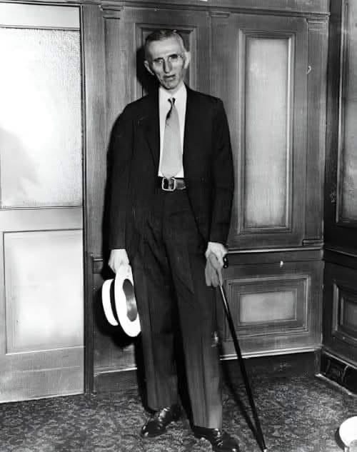 a black-and-white photo of a man in a suit holding a hat and cane, standing in front of wooden doors. The text overlaid on…