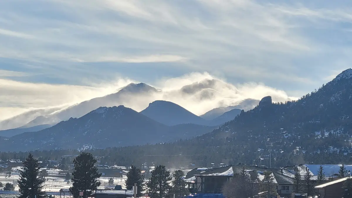A scenic winter landscape featuring snow-covered mountains partially obscured by clouds, with a residential area in the…