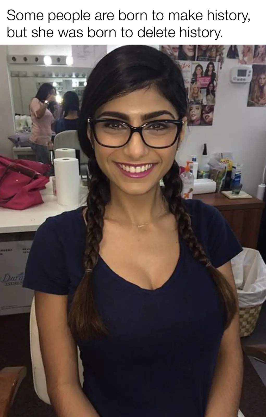 A woman with dark hair in two braids and wearing black-rimmed glasses smiles at the camera while seated in what appears to…