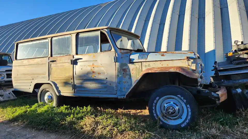 A heavily rusted, vintage, light-colored station wagon or van sits abandoned on grass in front of a large metal barn. The…