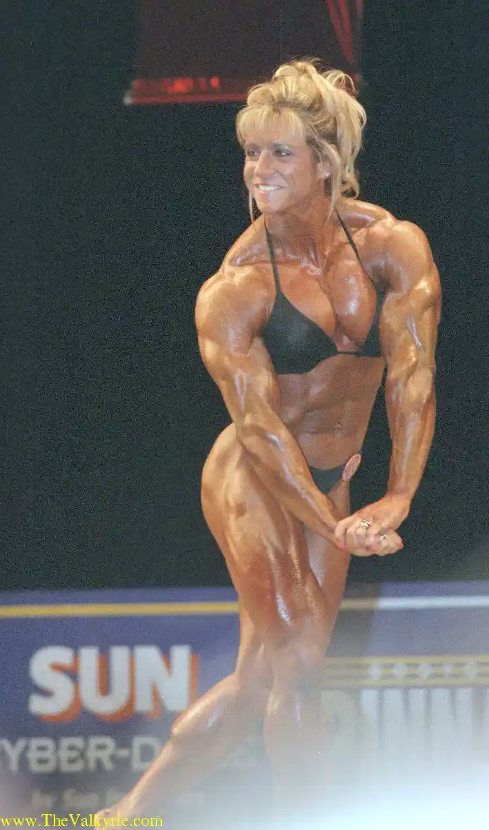 A muscular woman in a black bikini poses on a bodybuilding stage, her skin glistening with oil, showcasing extreme muscle…