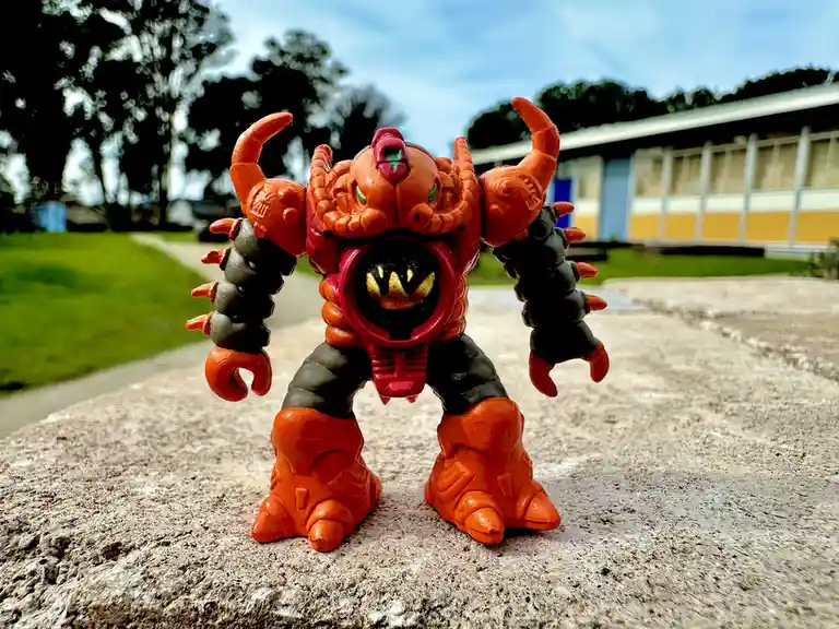 A small orange and black monster action figure with clawed arms and a circular chest plate stands on a concrete surface…