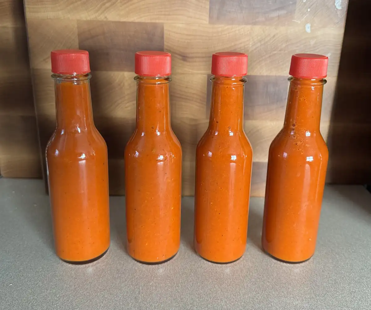 Four identical glass bottles filled with orange-red hot sauce and sealed with red caps are lined up on a countertop in front…