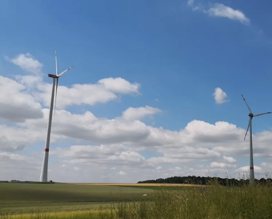 A scenic photo of two large white wind turbines standing in a green field under a blue sky with fluffy white clouds. The…