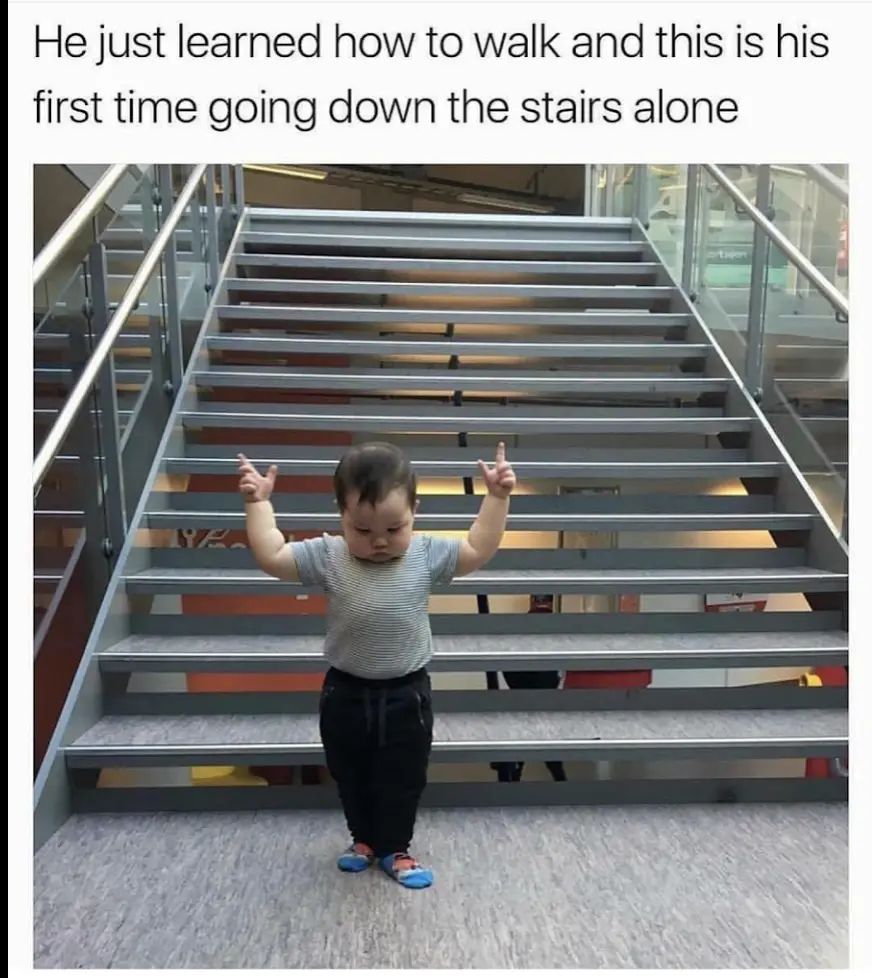A toddler in a striped shirt and dark pants stands at the bottom of a wide, modern metal staircase, arms raised with fingers…