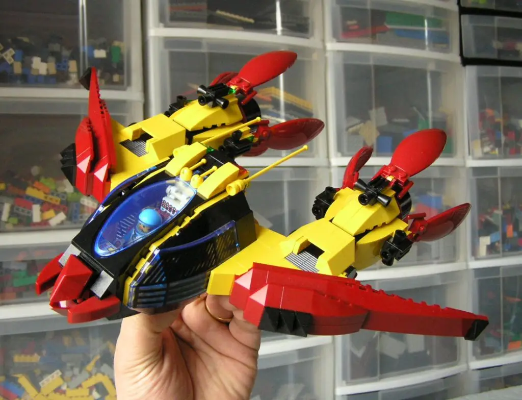 A hand holds a large, custom-built LEGO jet with a yellow body, red wings and tail fins, and a blue cockpit containing a…