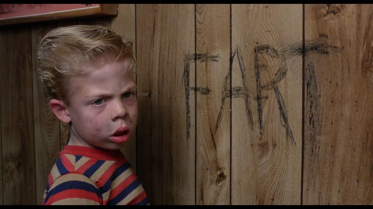 A young boy with blonde hair and a striped shirt leans against a wooden wall, looking over his shoulder with a disgusted…