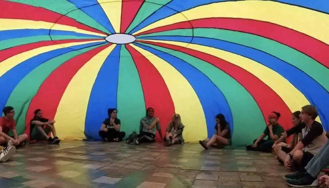 A group of people sit in a circle on a tiled floor beneath a large, colorful, rainbow-striped parachute canopy. The…
