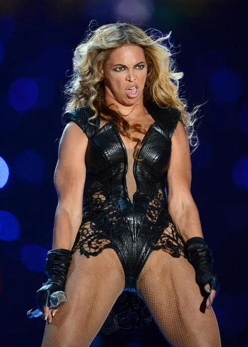 A woman with long blonde hair wearing a black leather and lace bodysuit with fishnet stockings and black gloves, holding a…