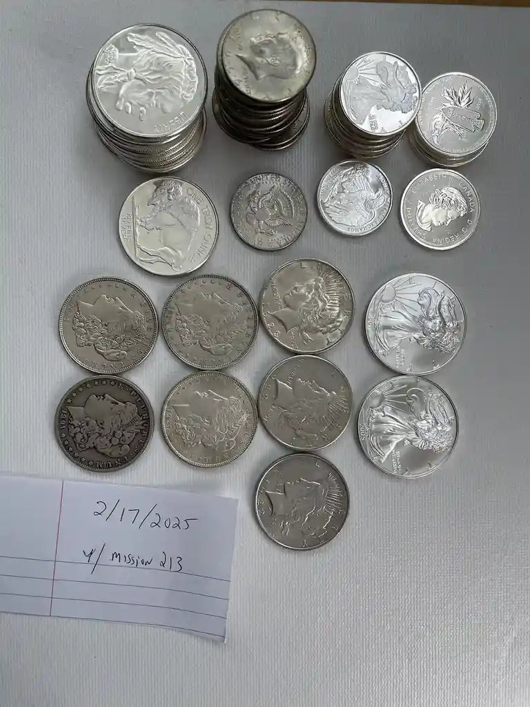 An overhead shot displays numerous silver coins of various designs and denominations arranged on a white textured surface…