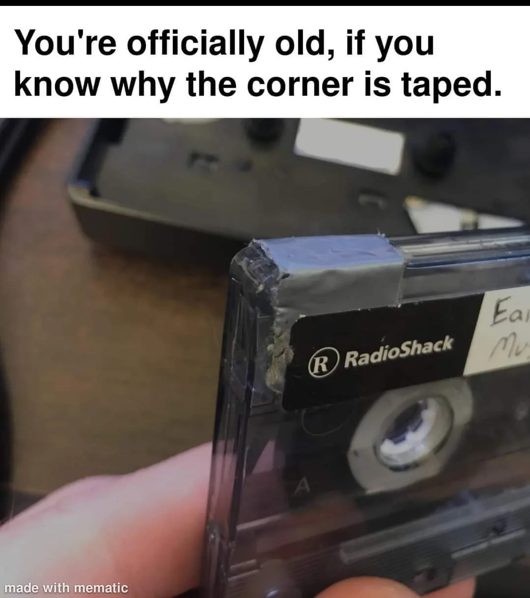 A close-up photo shows a hand holding a clear plastic RadioShack cassette tape with a piece of gray duct tape on its top…