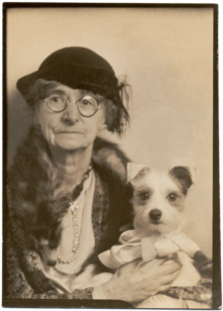 A sepia-toned vintage portrait shows an elderly woman wearing a hat and glasses, holding a small dog dressed in a white bow…
