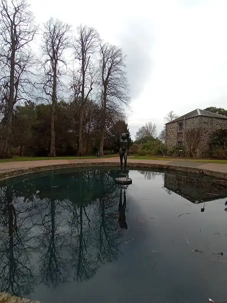 A bronze statue of a nude woman stands in the center of a calm, reflective pool, with bare trees and a stone building…