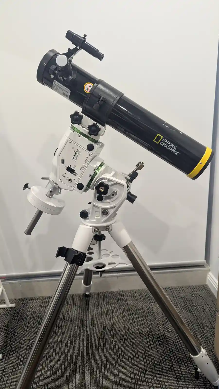 A black and white National Geographic branded telescope mounted on a tripod sits indoors on a carpeted floor against a white…