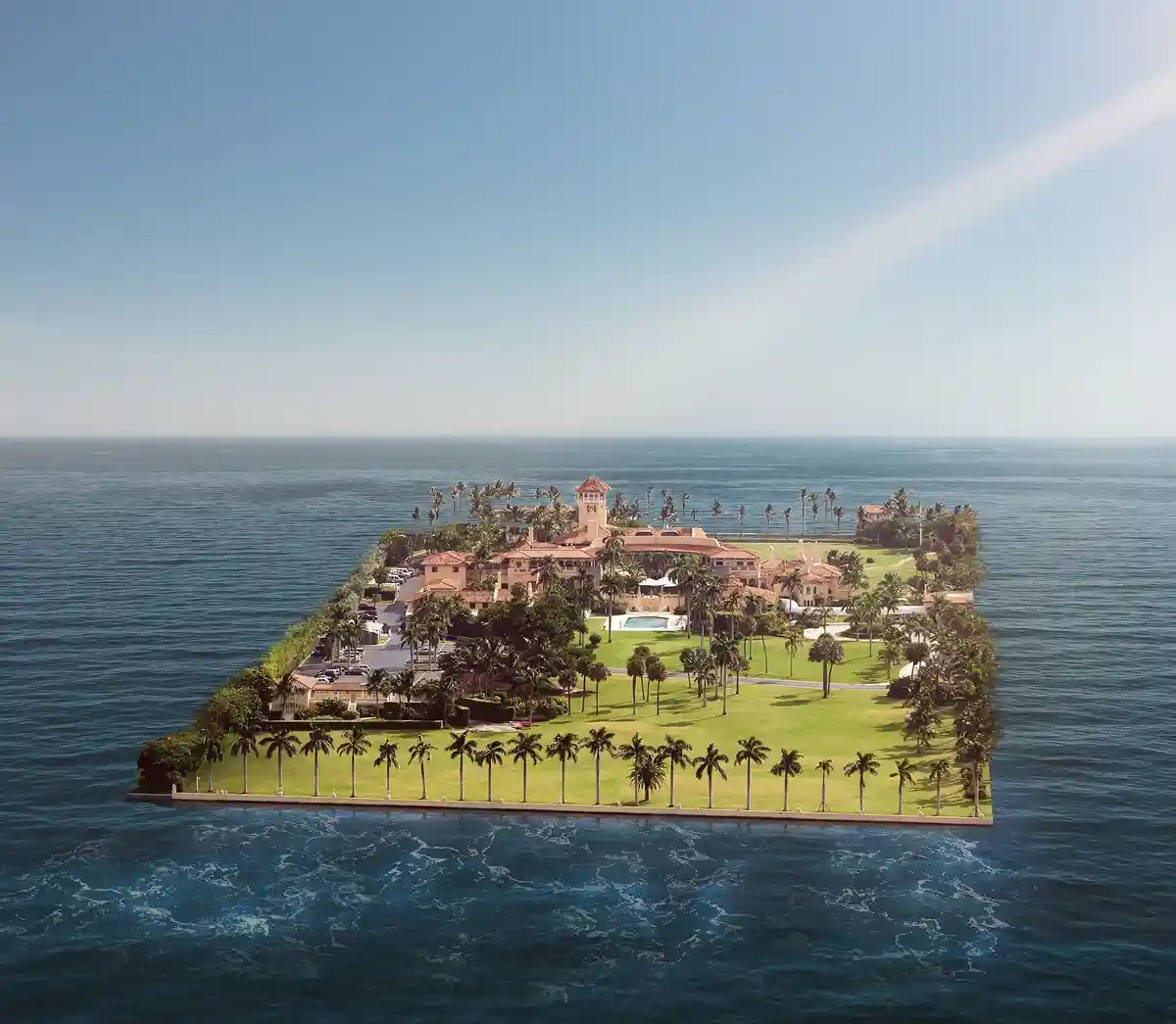 An aerial photograph shows a large, rectangular landmass surrounded by ocean, featuring a mansion, palm trees, manicured…